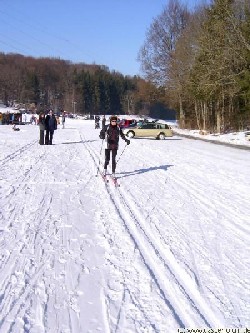 Eine Dame auf Langlaufski in der Loipe Engstingen unterwegs. Personen und Autos im Hintergrund.
